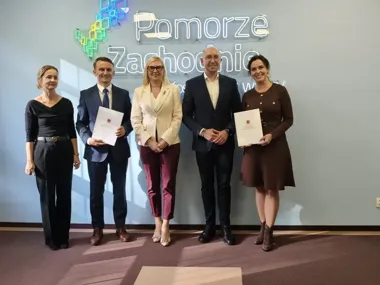 Pięć osób pozuje do zdjęcia przed neonowym napisem „Pomorze Zachodnie”, dwie z nich trzymają dokumenty.