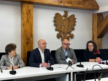 Cztery osoby siedzą przy stole z mikrofonami w pomieszczeniu z drewnianymi belkami i rzeźbionym orłem na ścianie.