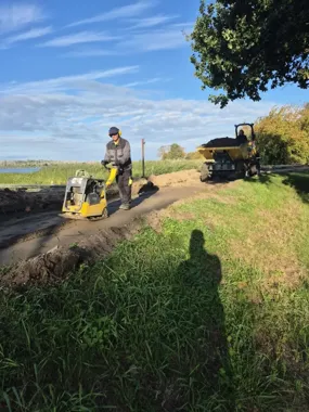 Robotnik w odzieży roboczej zagęszcza grunt za pomocą płyty wibracyjnej na drodze gruntowej, w tle stoi wywrotka z ładunkiem ziemi, a po lewej stronie widać jez
